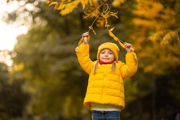 Gelukkig herfstconcept. een klein meisje in een gele jas en pet speelt met een boomtak en lacht. gelukkig meisje in de herfstpark. ruimte kopiëren.