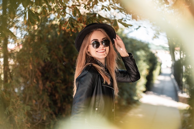 Gelukkig herfstportret van een mooie jonge vrouw met een glimlach in modieuze zwarte kleding met een hoed loopt buiten op een zonnige dag
