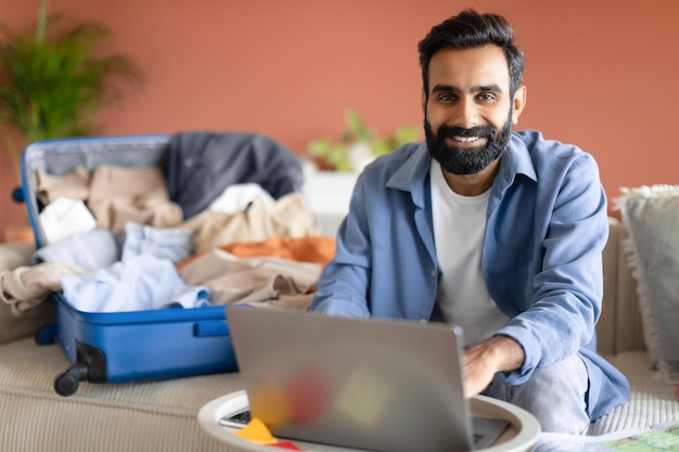 Gelukkig Indiase jonge man met behulp van laptop in de buurt van uitgepakte koffer binnen