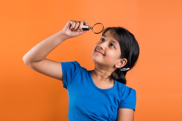 Gelukkig Indiase schoolmeisje kind met vergrootglas, staande geïsoleerd over oranje achtergrond