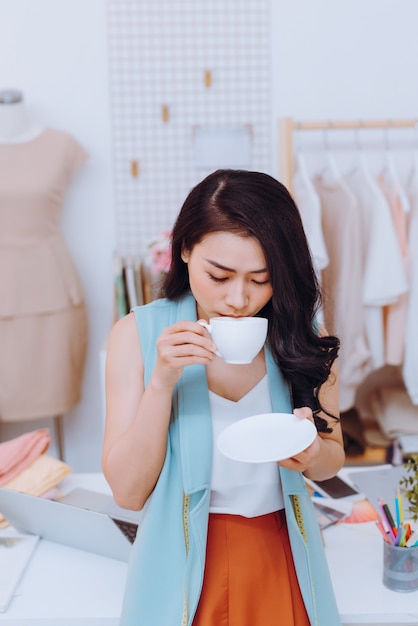 Gelukkig jong Aziatisch modeontwerperbedrijf dat koffie drinkt in haar werkplaats/winkel
