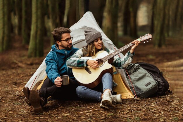 Gelukkig jong blank koppel in jassen in het bos geniet van een vakantie met een tent en speelt gitaar.