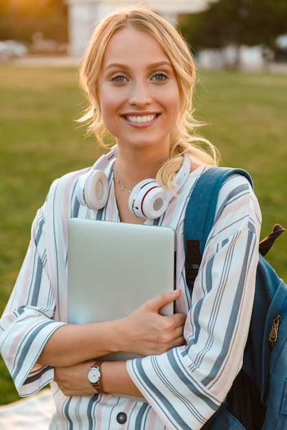 Foto gelukkig jong blond meisje met rugzak en laptopcomputer die in het park staat