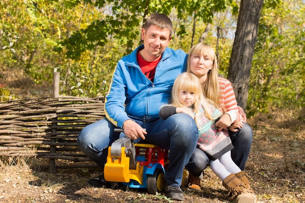 Gelukkig jong gezin met een moeder, vader en hun mooie blonde dochter poseren samen met een speelgoed dumper buiten in het bos in de zon tegen een rustieke houten hek