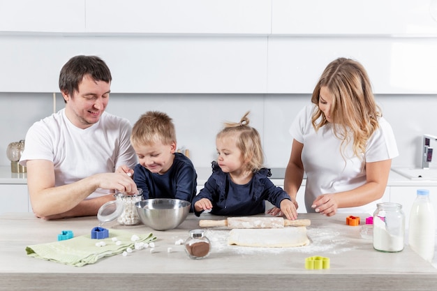 Gelukkig jong gezin met twee kleine kinderen bereidt deeg koekjes vrolijk in de keuken. Liefde en tederheid in een relatie.