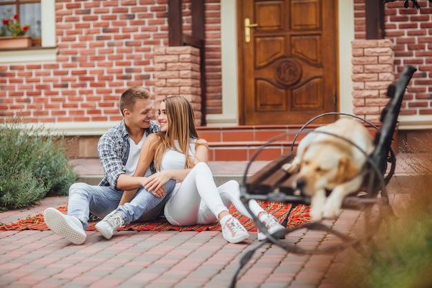 Foto gelukkig jong koppel genieten van elkaar en zitten aan het deken tapijt voor het huis. de hond slaapt op de bank