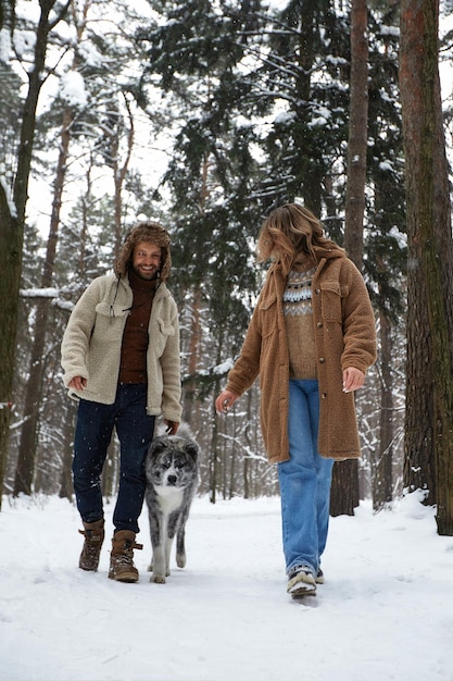 Gelukkig jong koppel in winterwear spelen met raszuivere Siberische husky en plezier in ingesneeuwd dennenbos