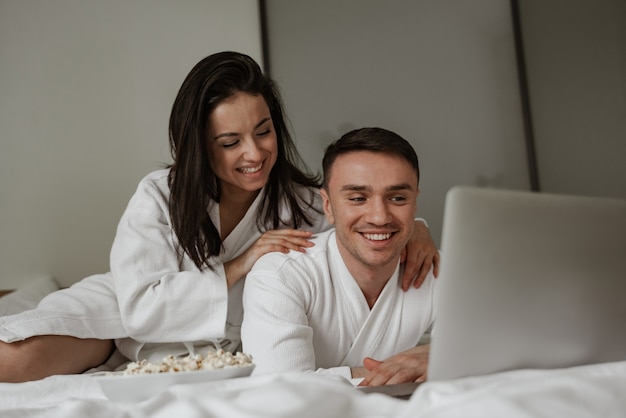 Gelukkig jong koppel in witte badjassen thuis in bed met popcorn, film kijken op laptop en lachen.