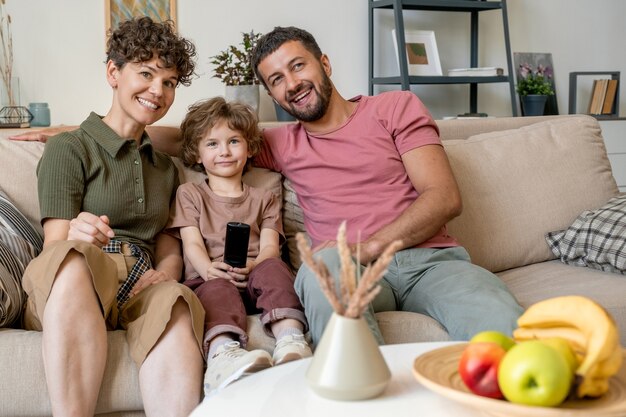 Gelukkig jong koppel lachen om tv-programma zittend op de Bank in de woonkamer met hun schattige zoontje tussen hen in