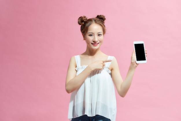 Gelukkig jong meisje gekleed in de zomer wijzende vinger op leeg scherm mobiele telefoon geïsoleerd over roze achtergrond