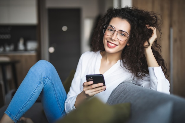 Gelukkig jong meisje het lezen van een bericht in een slimme telefoon zittend op een bank thuis