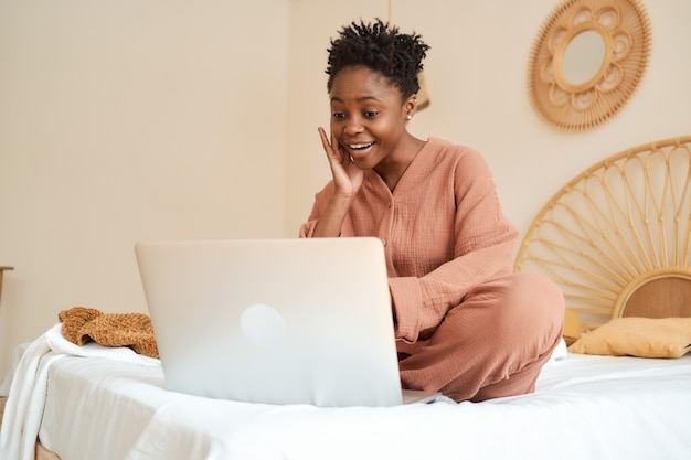 Gelukkig jong meisje zittend op bed in de slaapkamer en met behulp van laptop mousseline pyjama chatten met vrienden