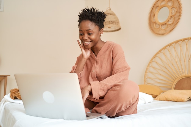 Gelukkig jong meisje zittend op bed in de slaapkamer en met behulp van laptop mousseline pyjama chatten met vrienden