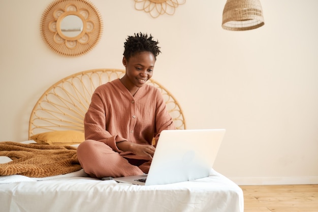 Gelukkig jong meisje zittend op bed in de slaapkamer en met behulp van laptop mousseline pyjama chatten met vrienden