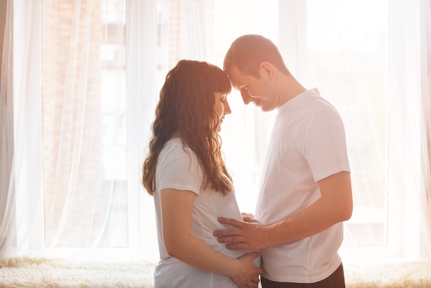 Gelukkig jong paar dat baby verwacht die zich samen voor venster bevindt dat een buik houdt en aan een baby denkt