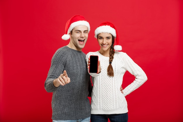 Gelukkig jong paar in Kerstmissweaters die vinger met mobiele telefoon richten.