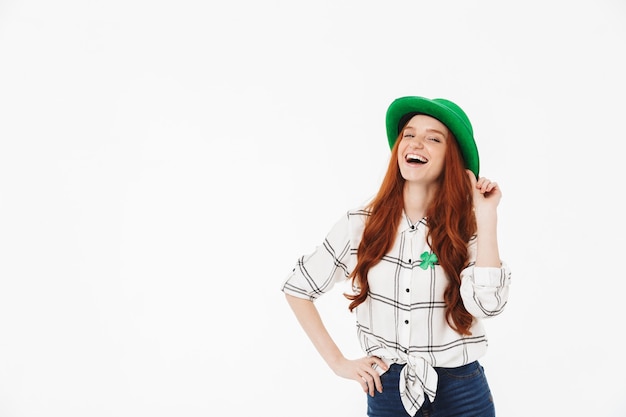 Gelukkig jong roodharige meisje met groene hoed, vieren St Patricks's Day geïsoleerd over witte muur, poseren