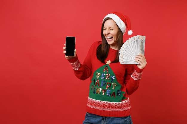 Gelukkig jong Santa meisje met veel stelletje dollars bankbiljetten, contant geld en mobiele telefoon met leeg leeg scherm geïsoleerd op rode achtergrond. Gelukkig Nieuwjaar 2019 viering vakantie partij concept.