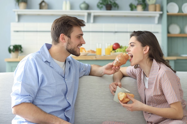 Gelukkig jong stel in pyjama's in de keuken aan het ontbijt, elkaar een croissant voeren.