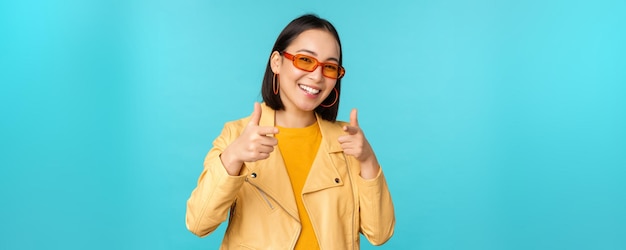 Gelukkig jong, stijlvol Chinees meisje in zonnebril wijst vingers naar de camera met een tevreden glimlach die kiest