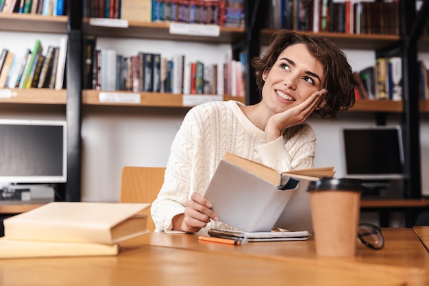 Gelukkig jong studentenmeisje dat bij de bibliotheek bestudeert, een boek leest