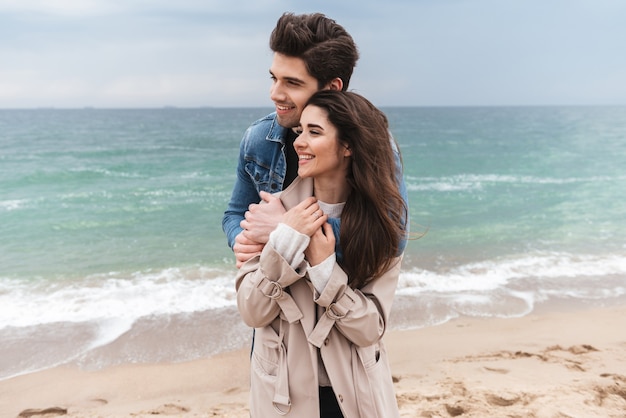 Gelukkig jong verliefd stel met herfstjassen die samen tijd doorbrengen aan de kust, knuffelen