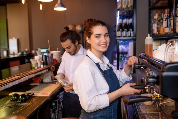 Gelukkig jonge aantrekkelijke serveerster van café of restaurant cappuccino bereiden door koffiemachine met collega op achtergrond