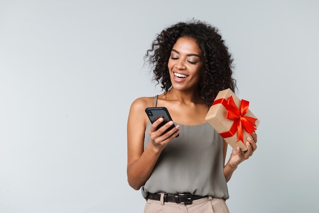 Gelukkig jonge Afrikaanse vrouw terloops gekleed staande geïsoleerd, met behulp van mobiele telefoon, met geschenkdoos