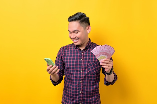 Gelukkig jonge Aziatische man met behulp van mobiele telefoon en het aanhouden van geld bankbiljetten geïsoleerd op gele achtergrond Fast credits concept