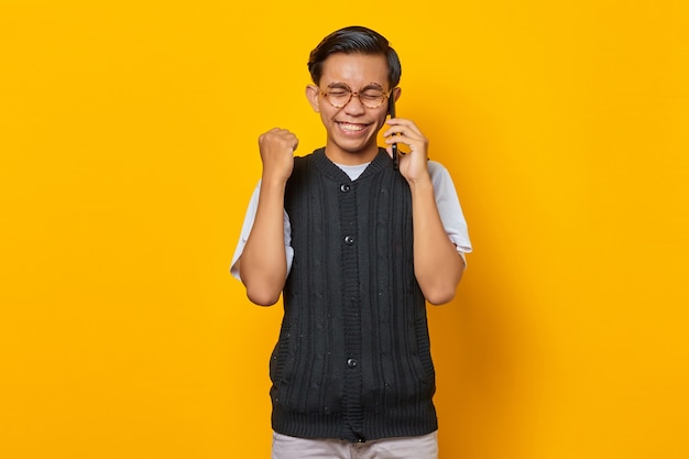 Gelukkig jonge Aziatische man met behulp van mobiele telefoon met handgebaar succes op gele achtergrond