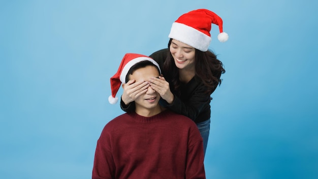 Gelukkig jonge Aziatische vrouw dragen van de Kerstman en bedek de ogen van een vriendje met haar hand geïsoleerd op blauwe achtergrond in studio opname. Kerstmis en gelukkig Nieuwjaar viering concept.