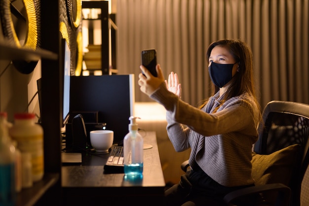 Gelukkig jonge Aziatische vrouw met masker videobellen tijdens het werken vanuit huis 's nachts