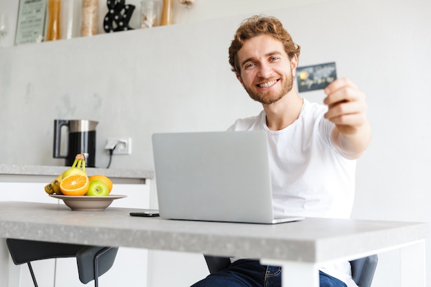 gelukkig jonge bebaarde man aan de tafel thuis praten met behulp van laptop computer bedrijf creditcard.