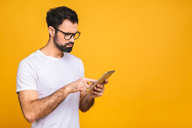 Gelukkig jonge bebaarde man in casual staande en met behulp van tablet geïsoleerd op gele achtergrond.