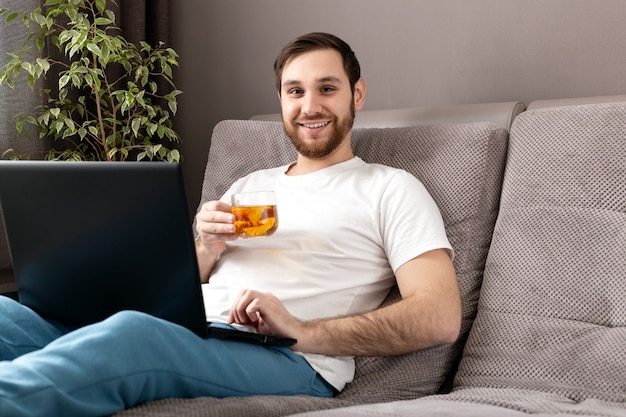 Gelukkig jonge blanke man drinkt thee tijdens het werken vanuit huis met behulp van laptop. Gezellig thuiskantoor,