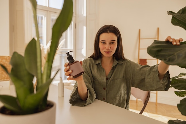 Gelukkig jonge blanke vrouw kijken camera met pot lotion zittend aan tafel binnenshuis brunette met lang haar draagt groen shirt schoonheid cosmetica en huidverzorging concept