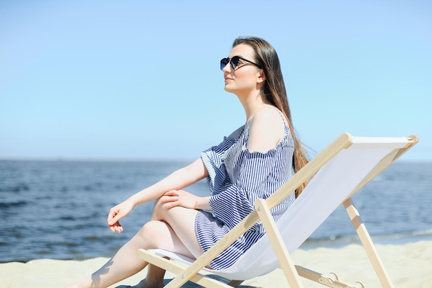 Gelukkig jonge brunette vrouw ontspannen op een houten ligstoel op het oceaanstrand terwijl ze glimlacht en een modezonnebril draagt. Het genieten van vakantie concept