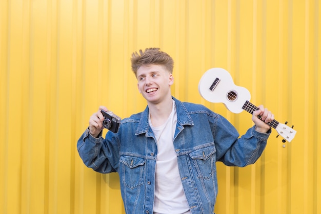 Gelukkig jonge hipster staat op een gele muur met ukelele en een retro camera in zijn handen