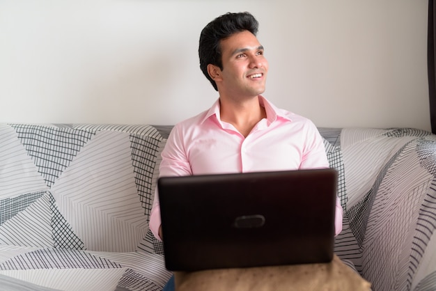 Gelukkig jonge indiase zakenman denken tijdens het gebruik van laptop thuis