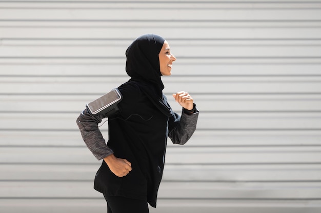 Gelukkig jonge islamitische vrouw atleet in hijab sportkleding met smartphone draait op muur achtergrond