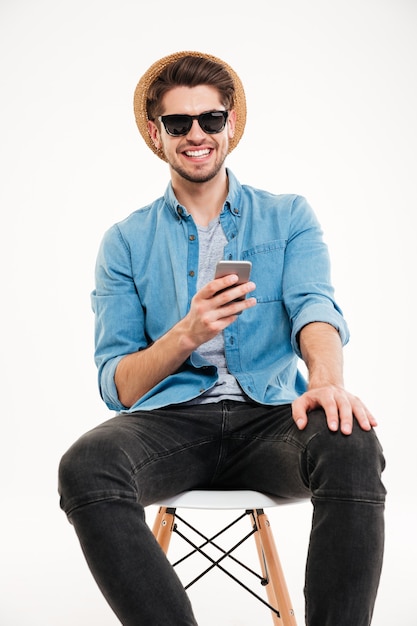 Gelukkig jonge man in blauw shirt, hoed en zonnebril met behulp van mobiele telefoon en glimlachen op witte achtergrond