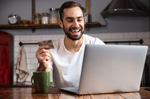 Gelukkig jonge man met laptopcomputer zittend aan de keukentafel, met creditcard