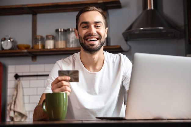 Gelukkig jonge man met laptopcomputer zittend aan de keukentafel, met creditcard