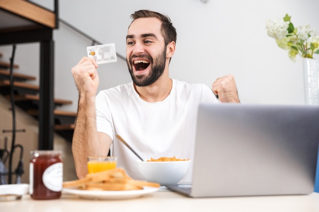 Gelukkig jonge man ontbijten in de keuken, met behulp van laptop computer, creditcard weergegeven: