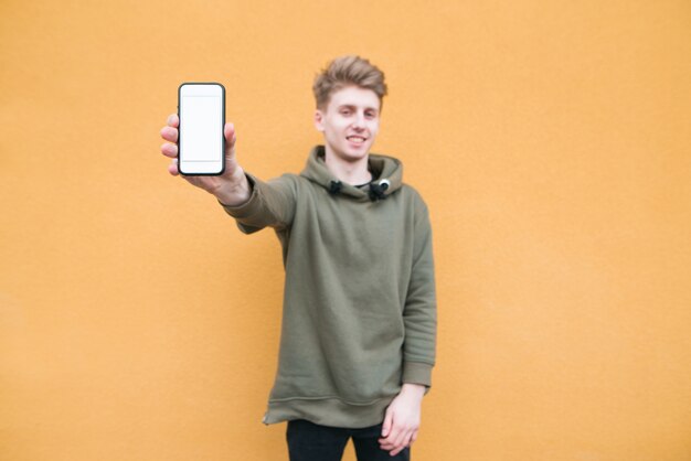 Gelukkig jonge man staat op een oranje muur, toont een smartphone met een wit scherm
