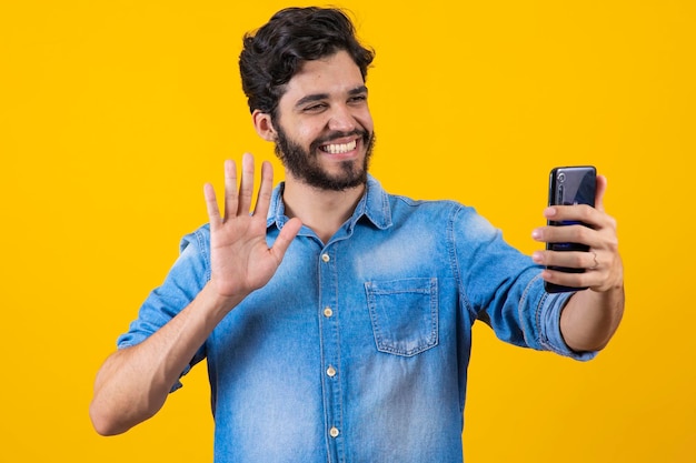 Gelukkig jonge man videobellen online praten met mobiele telefoon hallo zeggen tegen smartphone camera en hand vriendelijk zwaaien over gele achtergrond