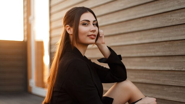 Gelukkig jonge mode vrouw in een zwarte jas zit in de buurt van een houten vintage muur bij zonsondergang