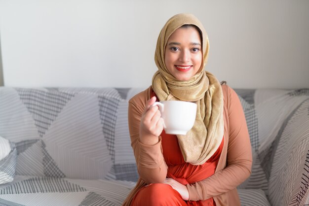 Gelukkig jonge mooie moslimvrouw met koffie in de woonkamer thuis