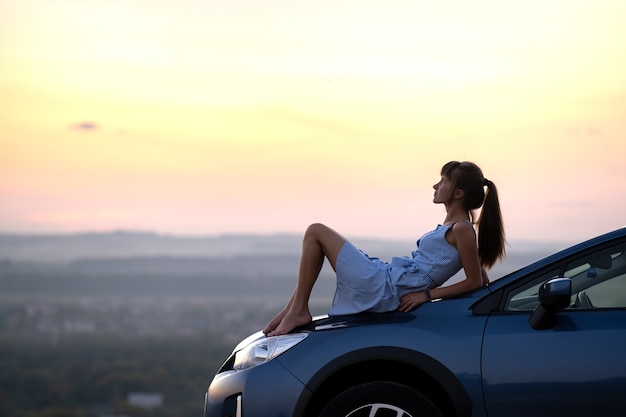 Gelukkig jonge vrouw bestuurder in blauwe jurk genieten van warme zomeravond liggend op de motorkap van haar auto. Reizen en vakantie concept.