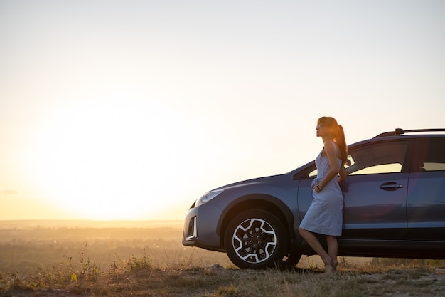 Gelukkig jonge vrouw bestuurder in blauwe jurk leunend op haar auto genieten van warme zomerdag. Reizen en vakantie concept.